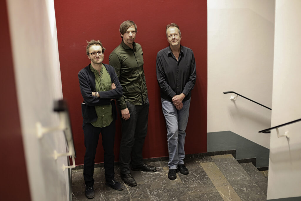 Ein Foto der drei Musiker Peter Ehwald, Stefan Schultze und Tom Rainey. Sie stehen in einem Treppenhaus vor einer dunkelroten Wand. Alle drei blicken in die Kamera.